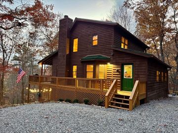 Dark wooden cabin with lit windows and porch in a gravel clearing among autumn trees at dusk