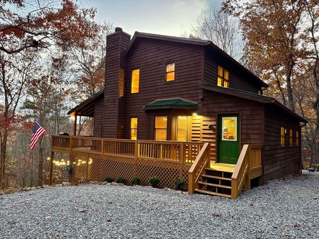 Dark wooden cabin with lit windows and porch in a gravel clearing among autumn trees at dusk