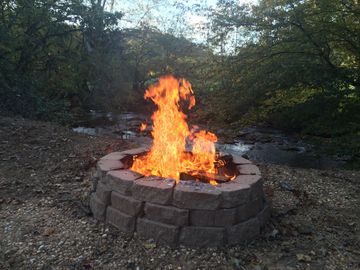 A bright campfire burns in a circular stone fire pit in a wooded outdoor setting.