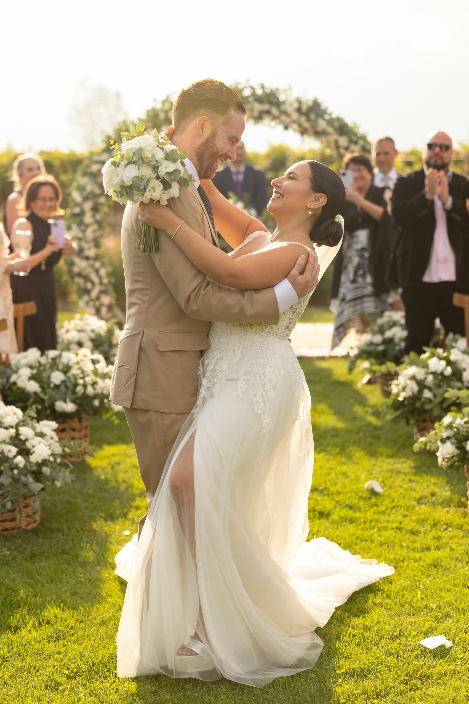Wedding couple dancing outdoors on a sunlit lawn, surrounded by white flowers and guests.