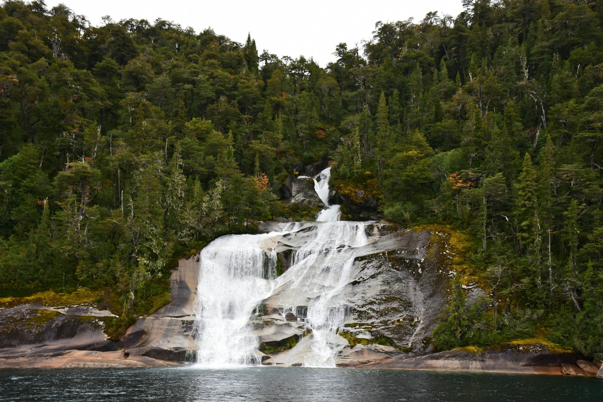 Waterfall cascading over rocky ledges in a dense green forest beside a calm river