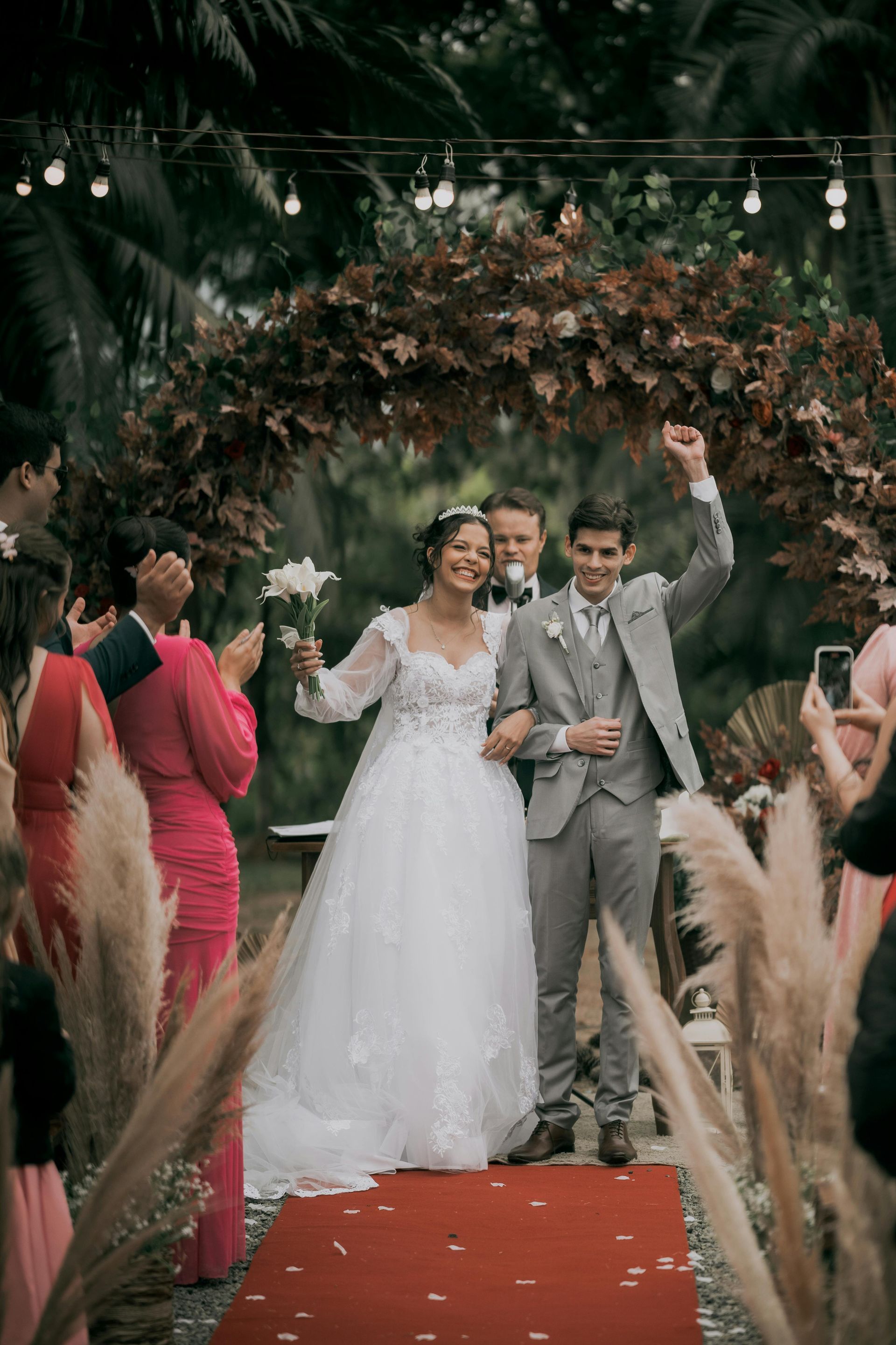 Newlyweds walk down a red aisle under a floral arch, smiling and waving at guests.