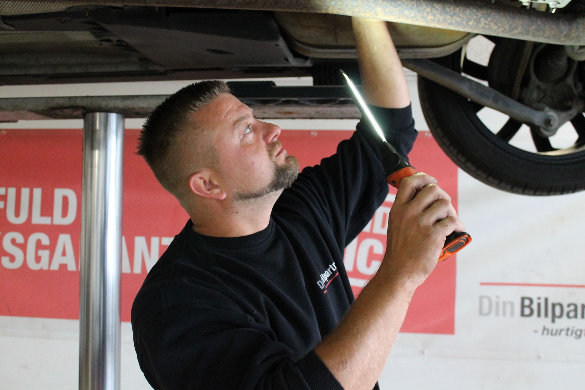 Mekaniker inspicerer undersiden af ​​en bil med en lommelygte. Han kigger op under bilen på en lift.