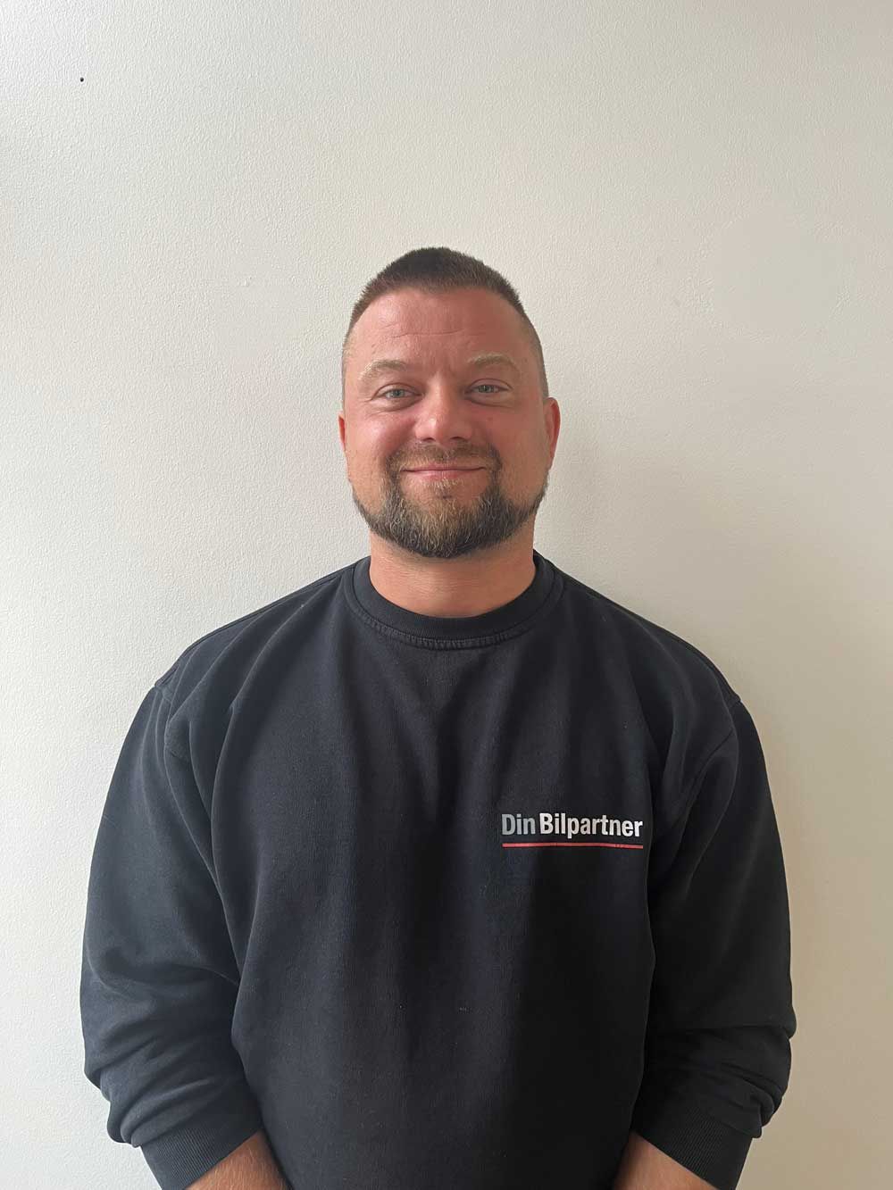 Mand med kort hår og skæg iført en sort sweatshirt med logo, smilende foran en hvid væg.