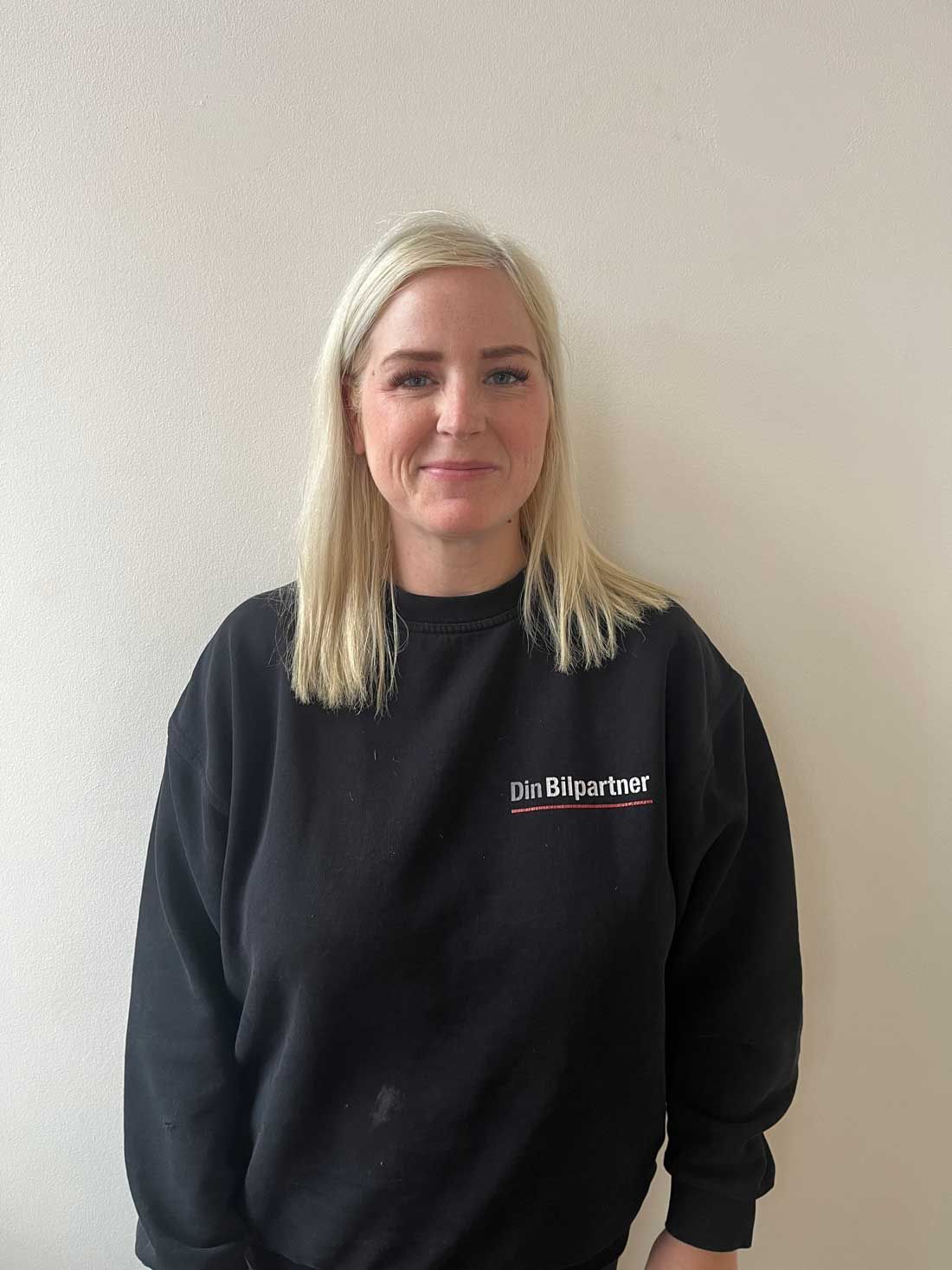 Blond kvinde i sort sweatshirt står op ad en hvid væg; smilende og kigger ind i kameraet.
