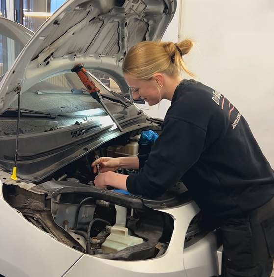 Kvinde i sort sweatshirt arbejder på en bilmotor i en garage; motorhjelmen er åben.