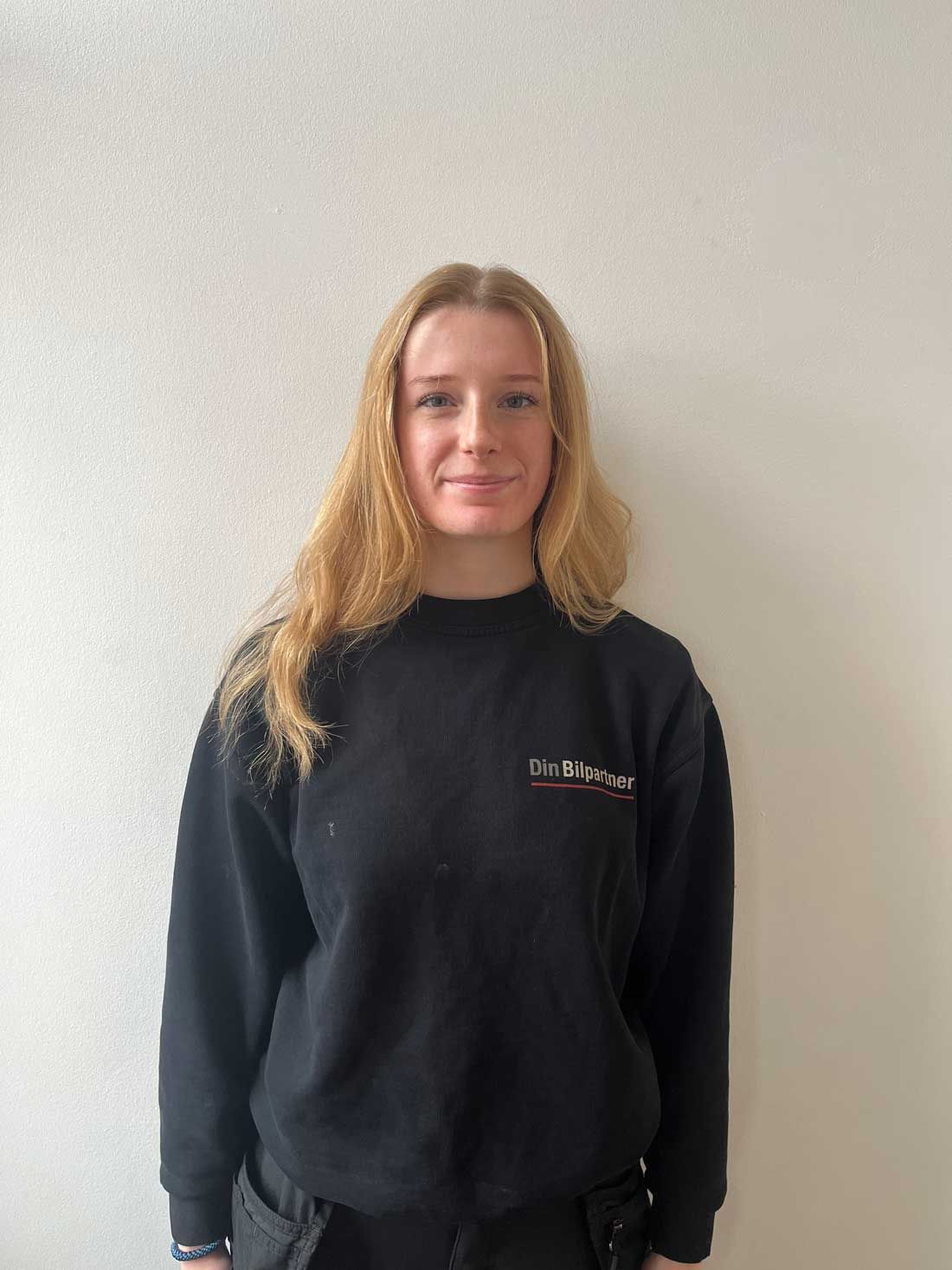 Kvinde i sort sweatshirt smilende, stående op ad en hvid væg.
