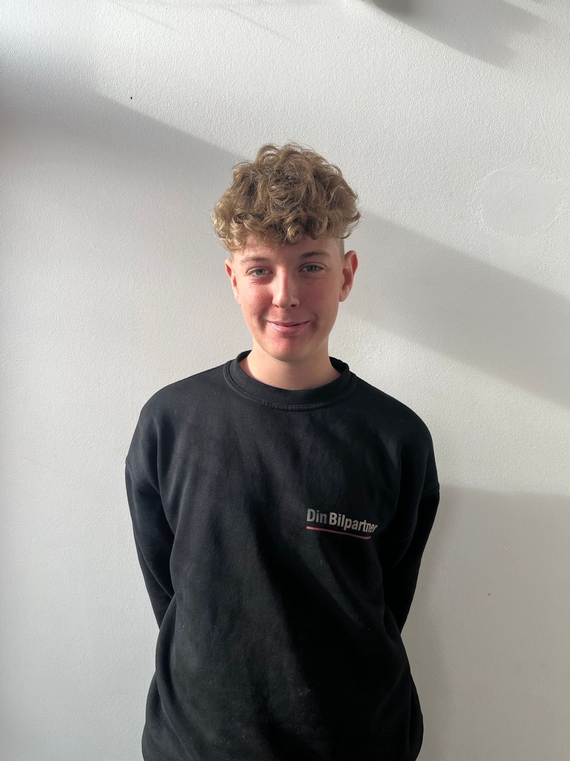 Ung person med krøllet hår iført en sort sweater, stående op ad en hvid væg.