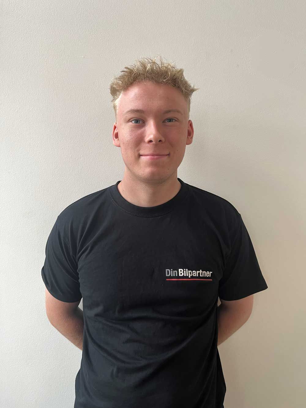 Mand i sort t-shirt med logo, hænderne på ryggen, smilende let, op mod en hvid væg.