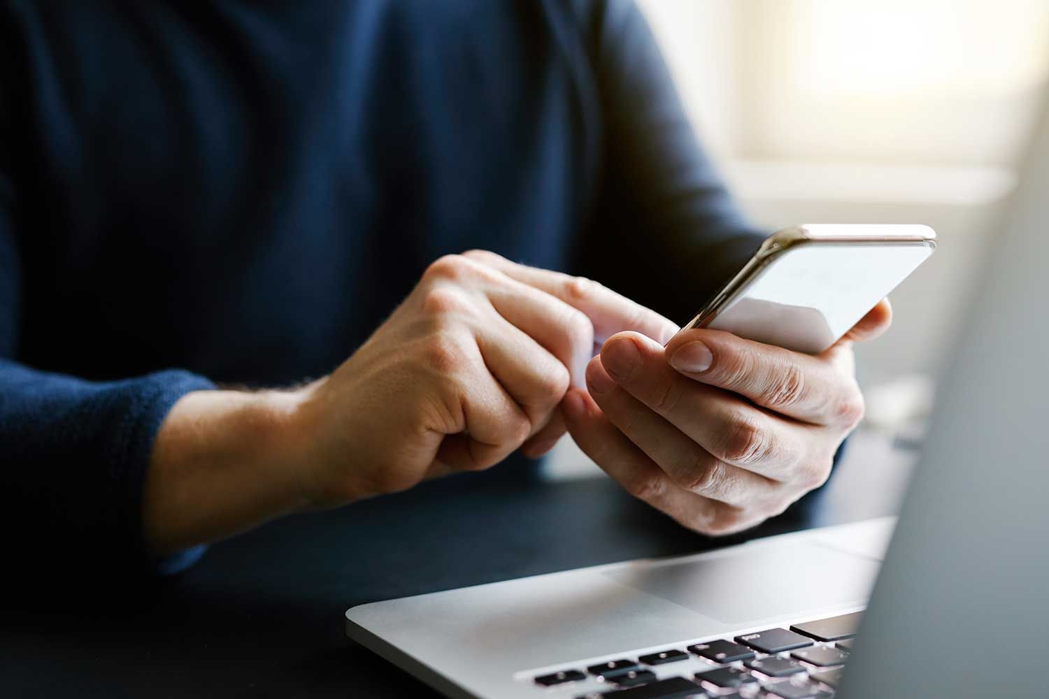 Person, der bruger en smartphone med en bærbar computer i forgrunden, indendørs.