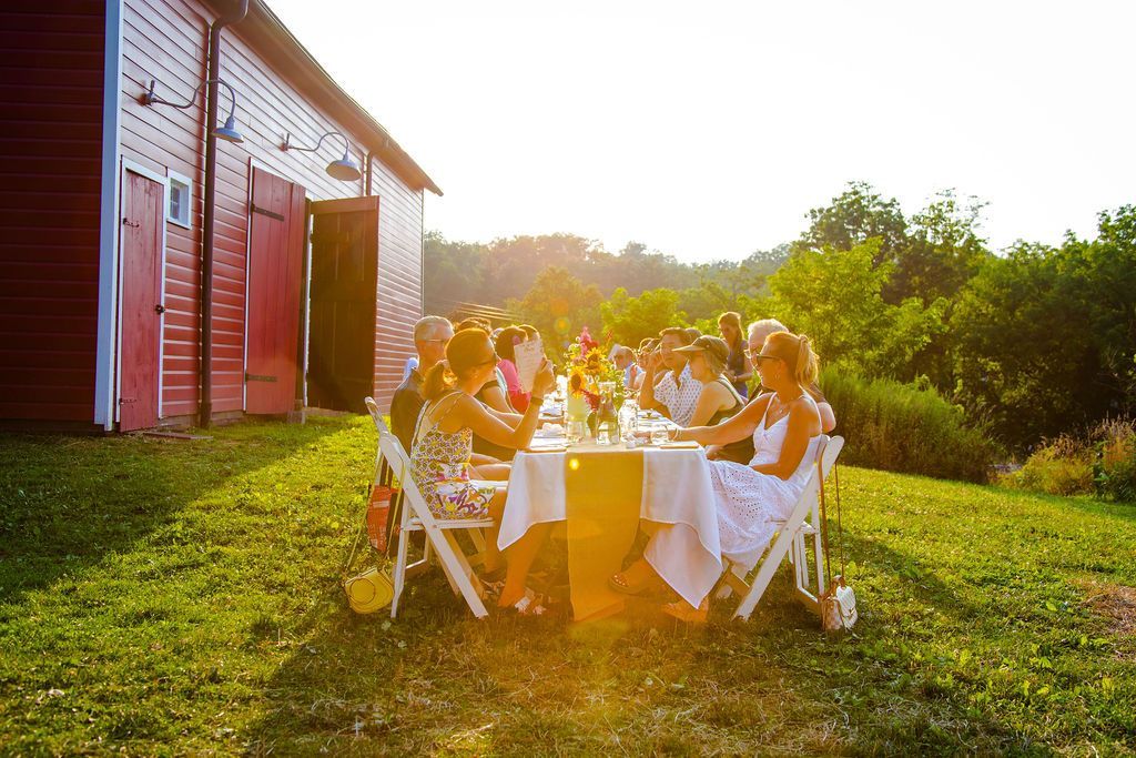 People seated at a long table, set for a meal, in front of a red barn. Golden light bathes the scene.