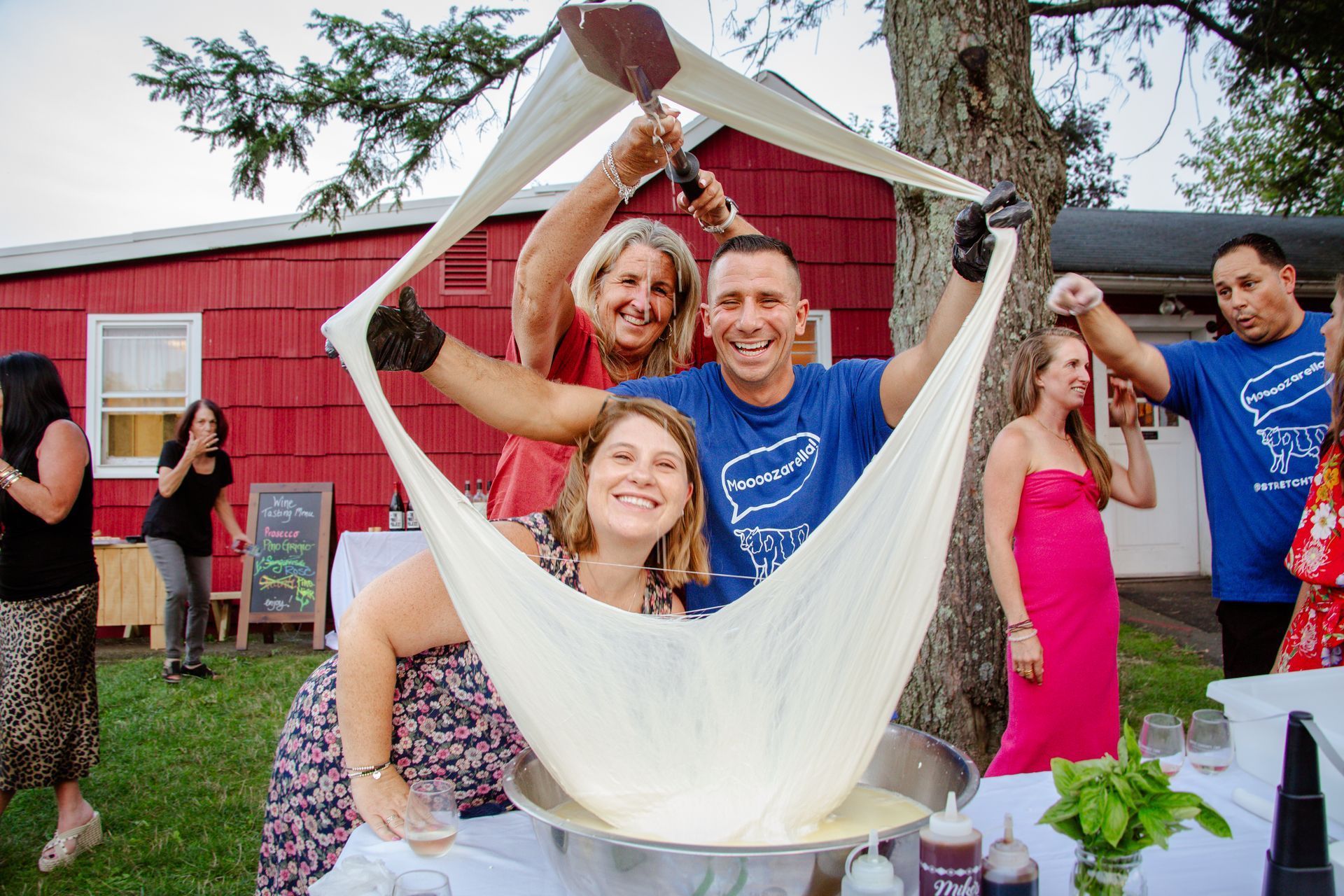 People stretching cheese at an outdoor event near a red building.