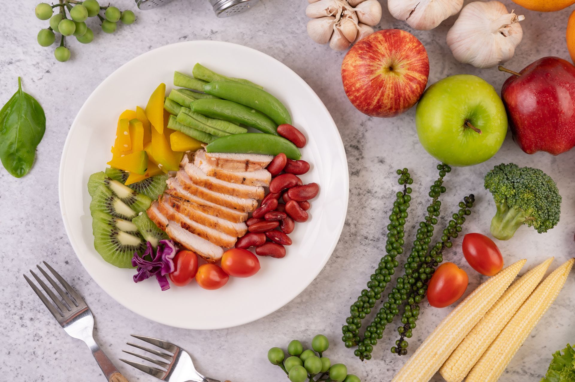 A top-down view of a plate with grilled chicken, kiwi, yellow squash, green beans, and kidney beans, near fresh produce.