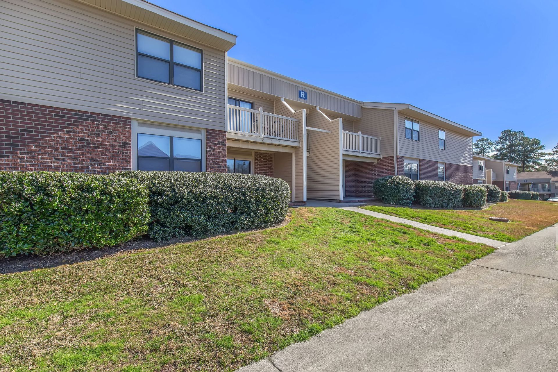 A two-story apartment building with brick and beige siding, featuring private balconies and manicured hedges.