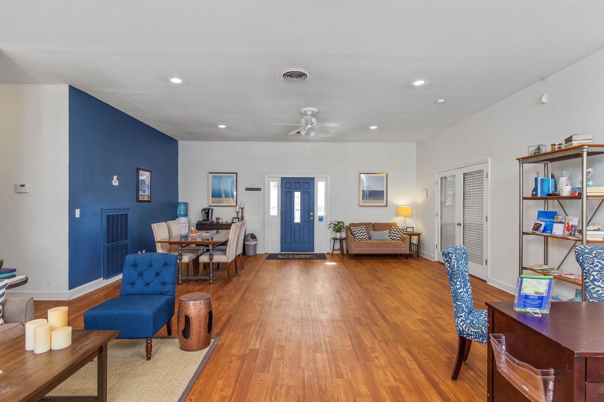 A spacious room with wood floors, a blue accent wall, a blue armchair, a dining area, and a beige sofa near the entrance.