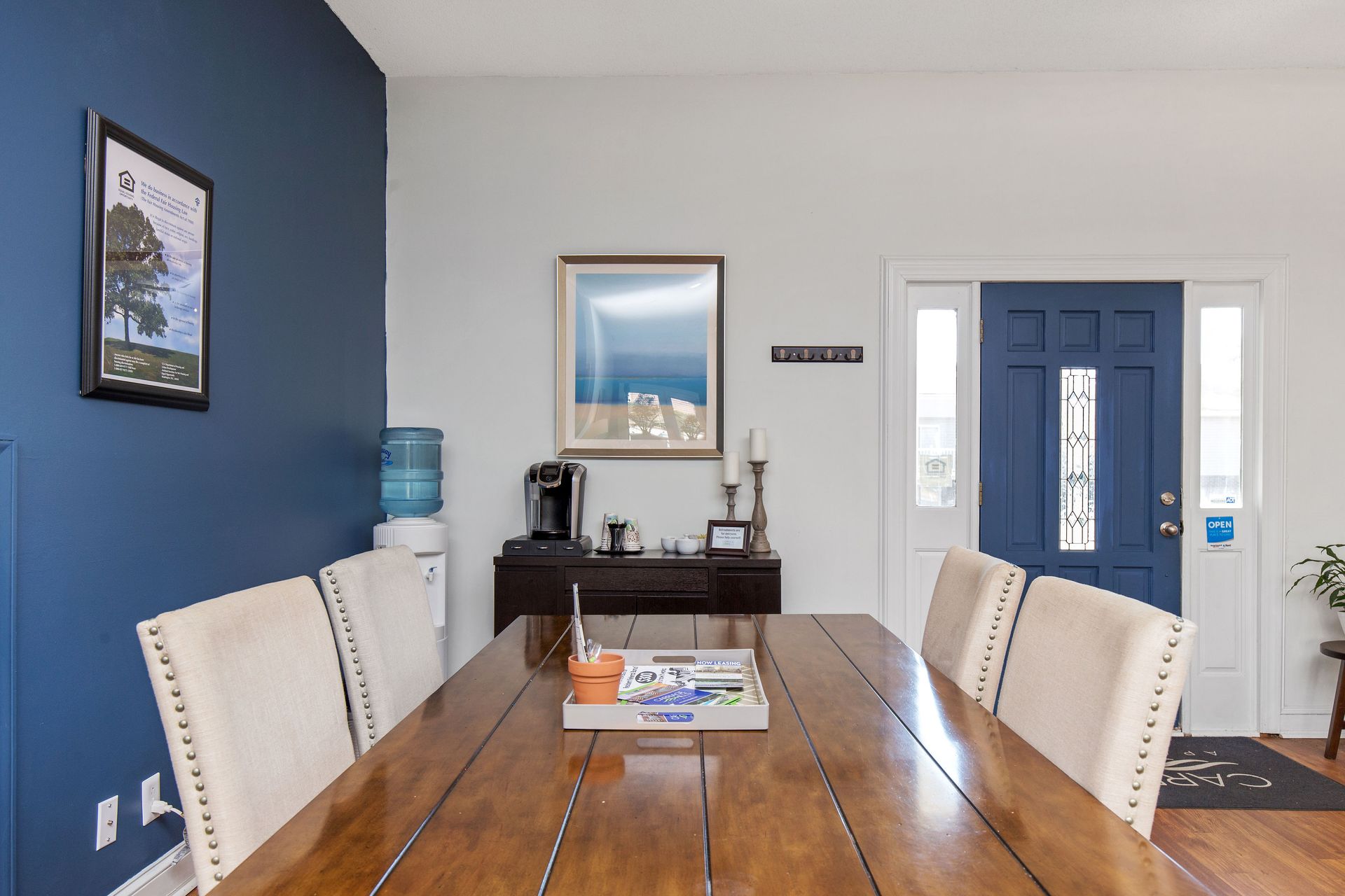 Dining area with a wooden table, four upholstered chairs, a navy accent wall, and a blue front door.