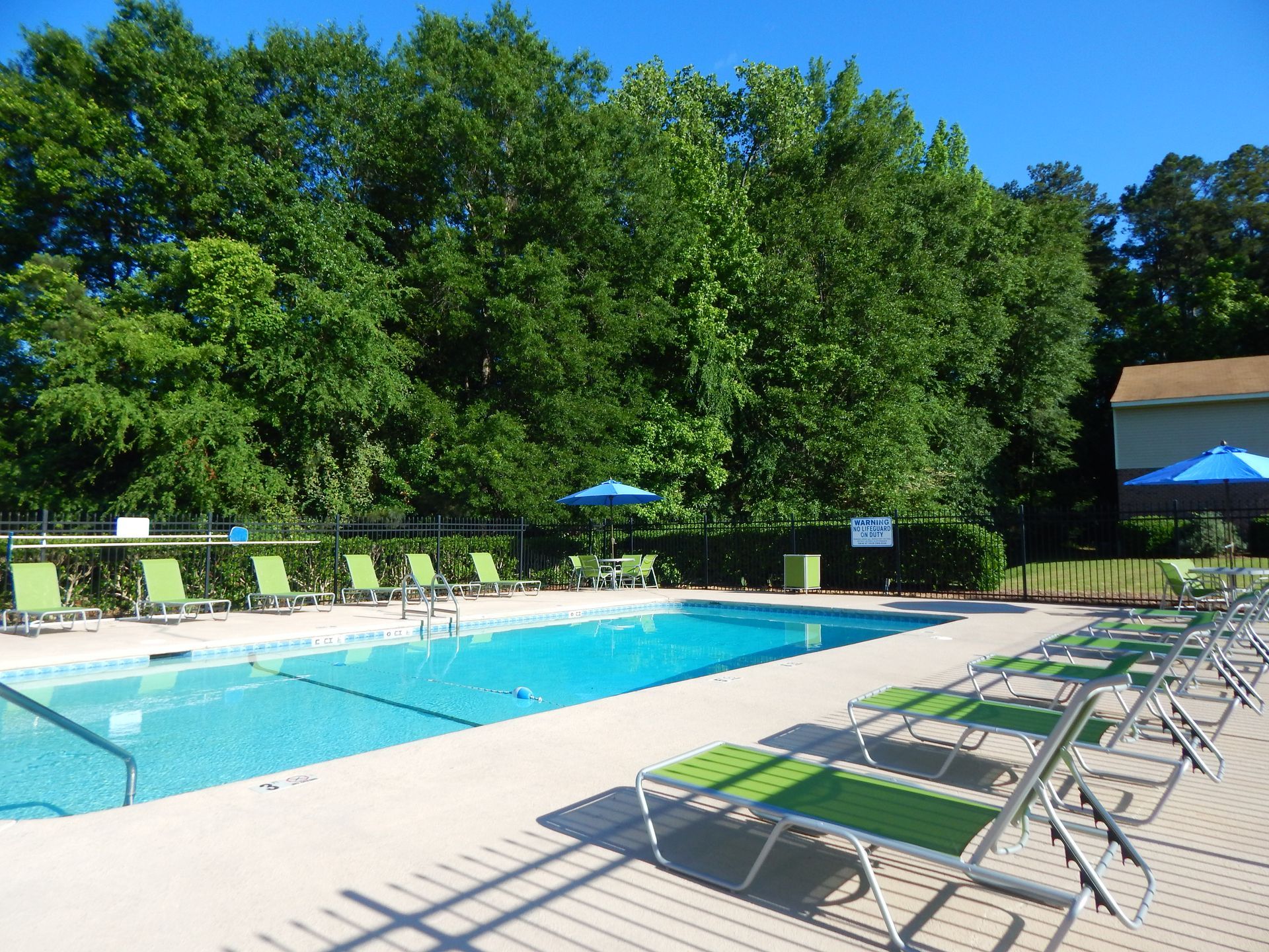 An outdoor rectangular swimming pool with bright green lounge chairs and blue umbrellas, set against a backdrop of trees.