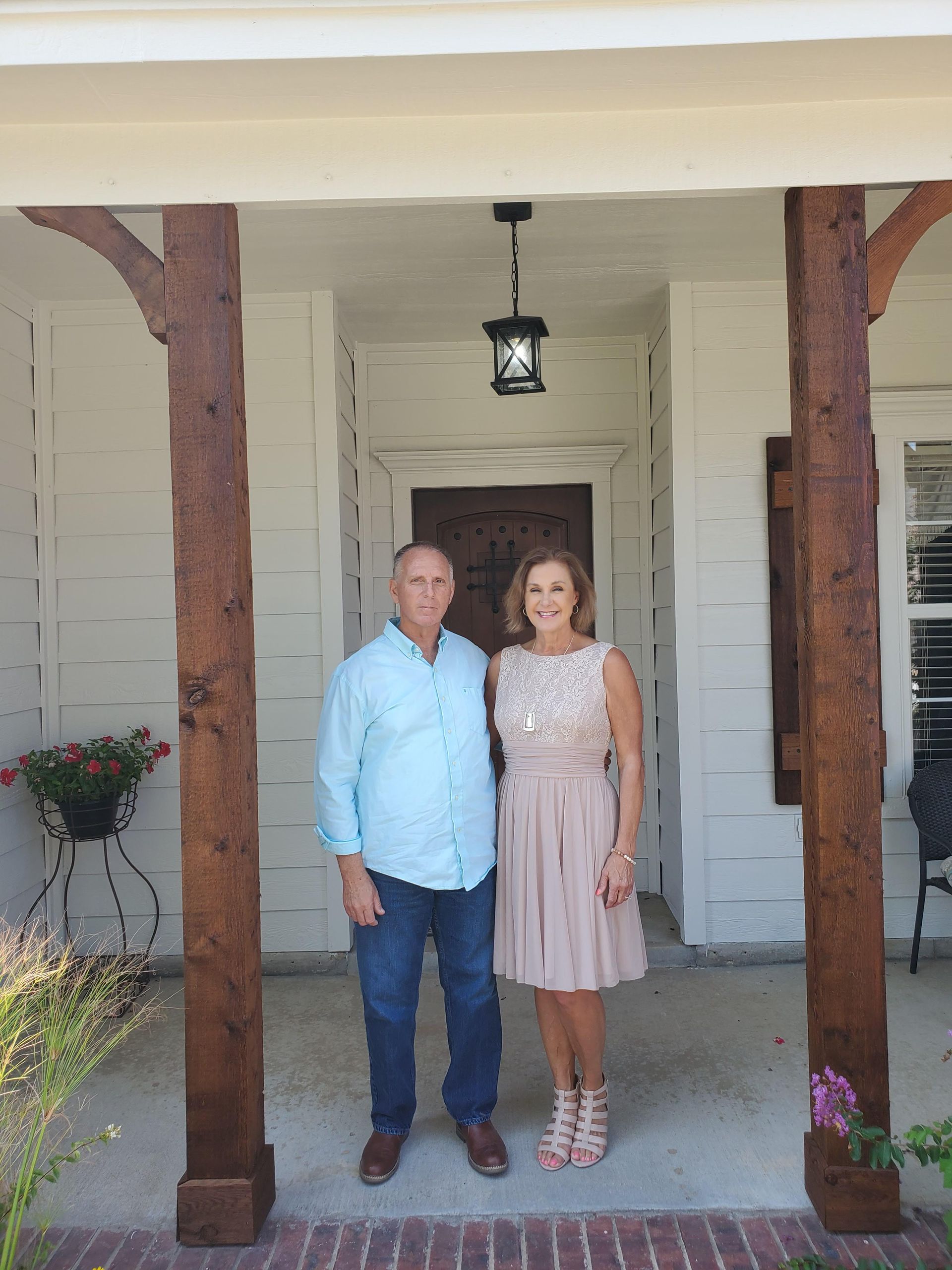 Couple standing in front of a white house with brown support beams. The woman is in a pink dress, and the man is in a blue shirt.