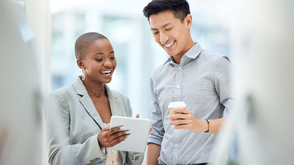 Mulher e homem olhando juntos para um tablet, sorrindo. O homem segura uma xícara de café. Ambiente de escritório.