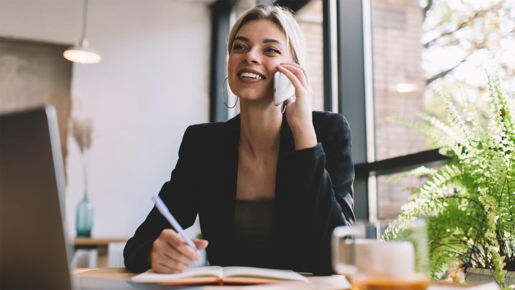 Mulher de blazer ao telefone, fazendo anotações em uma mesa perto de uma janela.