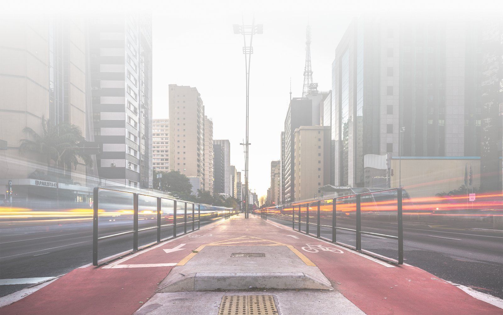 Rua da cidade com ciclovia vermelha, prédios altos e rastros de luz dos carros que passam.