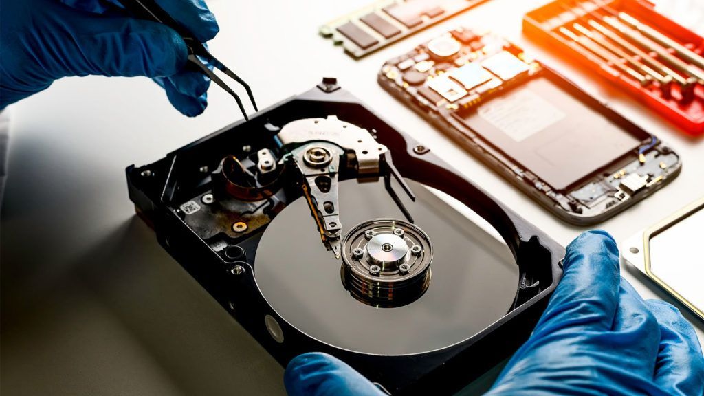 Pessoa com luvas azuis usando pinças em um disco rígido desmontado; um telefone desmontado e peças estão ao fundo.