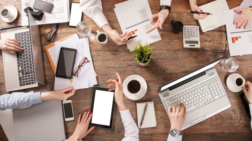 Mãos trabalhando em laptops, tablets e documentos ao redor de uma mesa de madeira em uma reunião de negócios.