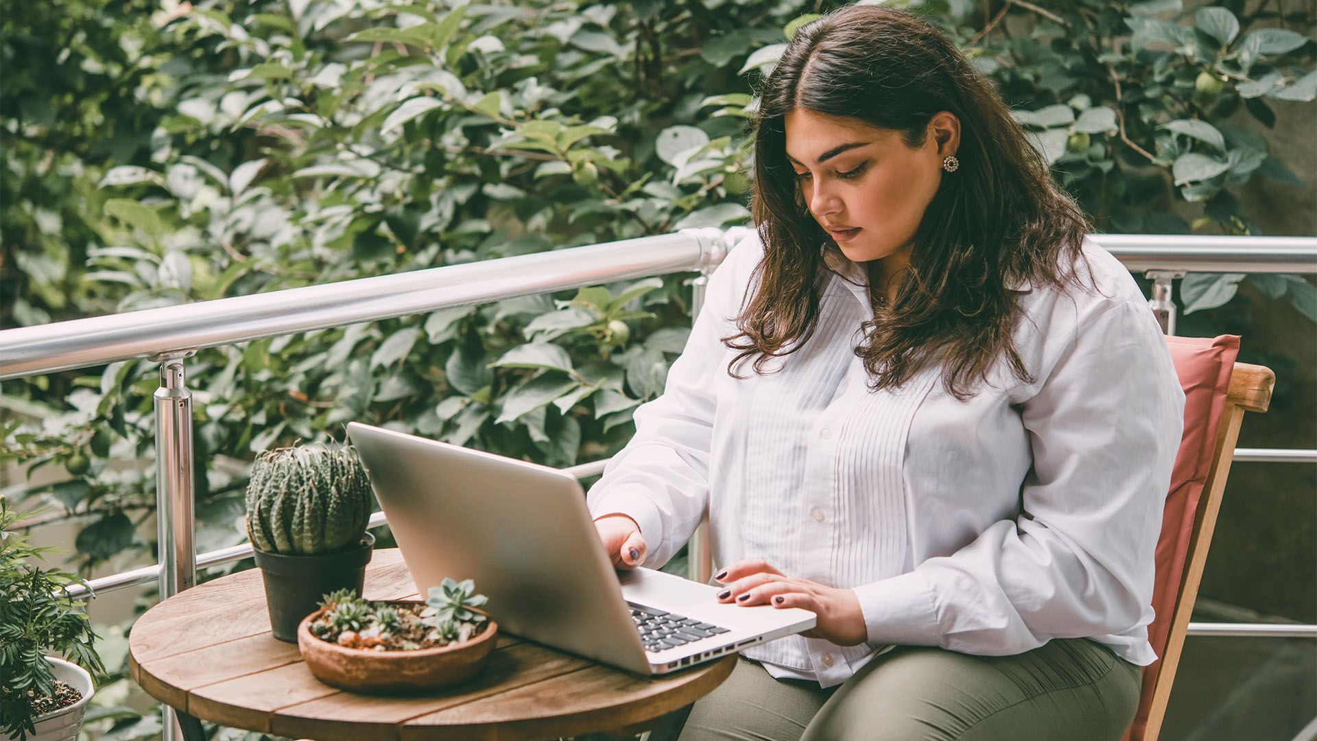 Mulher em uma varanda, usando um laptop, com plantas em uma pequena mesa.
