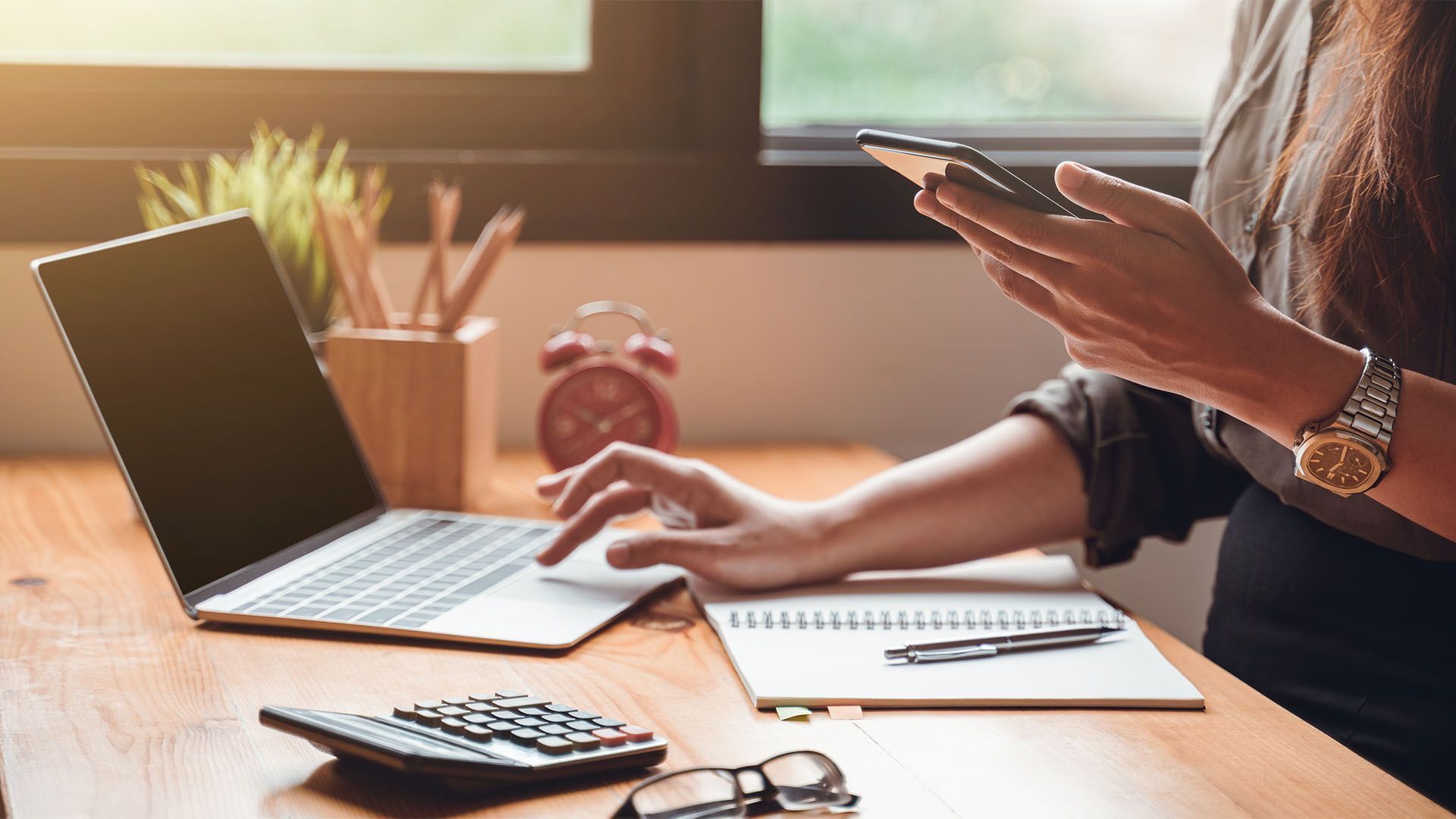 Mulher sentada à mesa usando laptop e celular, com bloco de notas, calculadora e relógio.