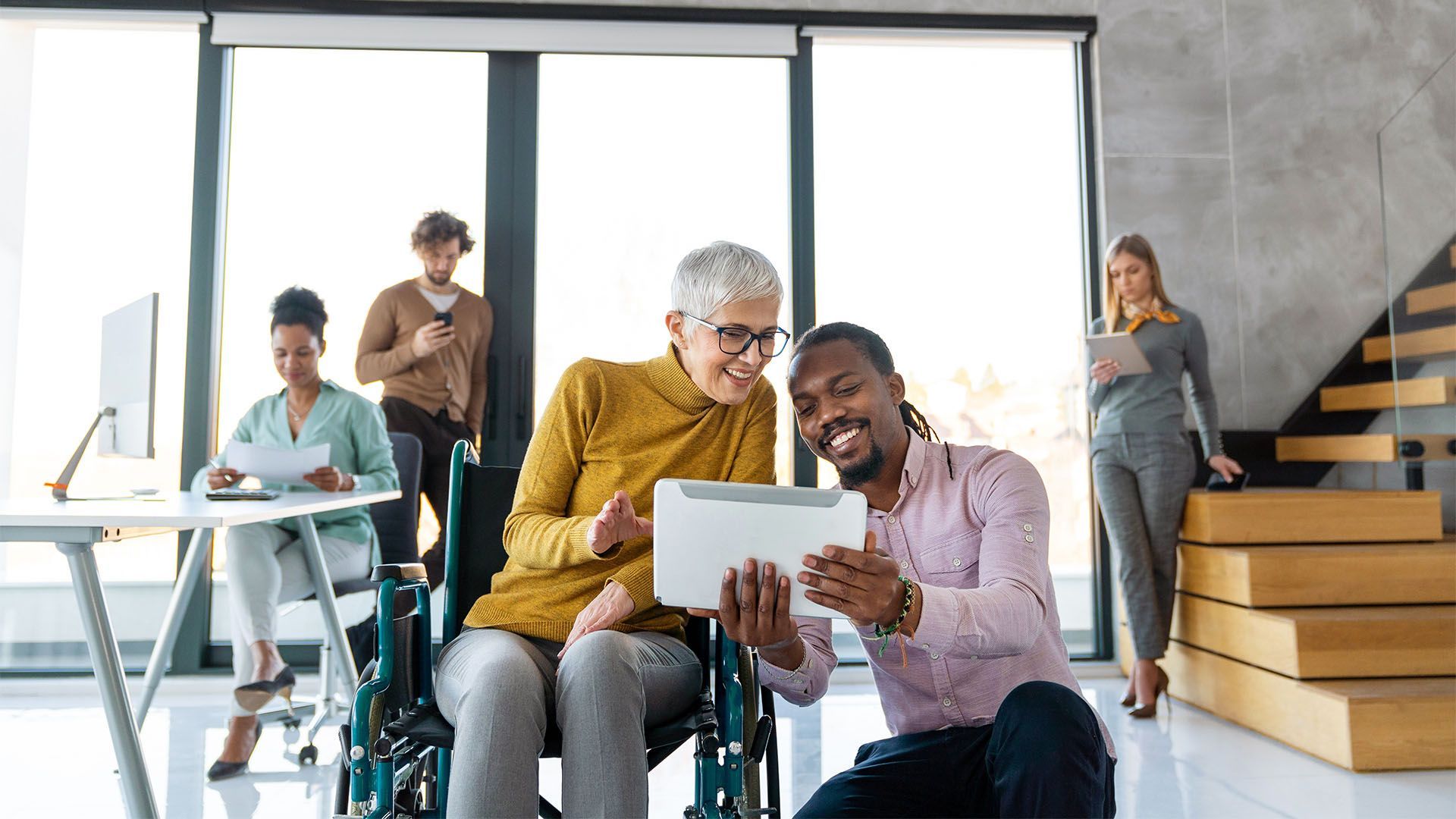 Diversos funcionários de escritório olhando juntos para um tablet, mulher em cadeira de rodas. Luz solar entrando pelas grandes janelas.