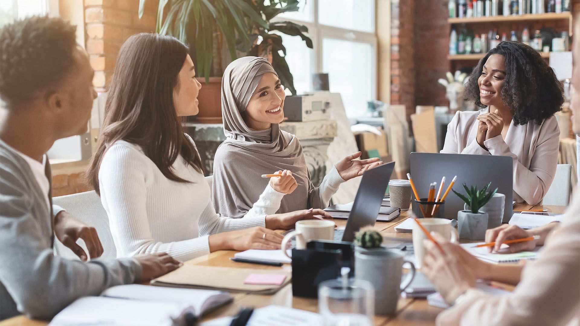 Grupo de pessoas reunidas em torno de uma mesa com laptops, conversando e sorrindo em um ambiente de escritório.