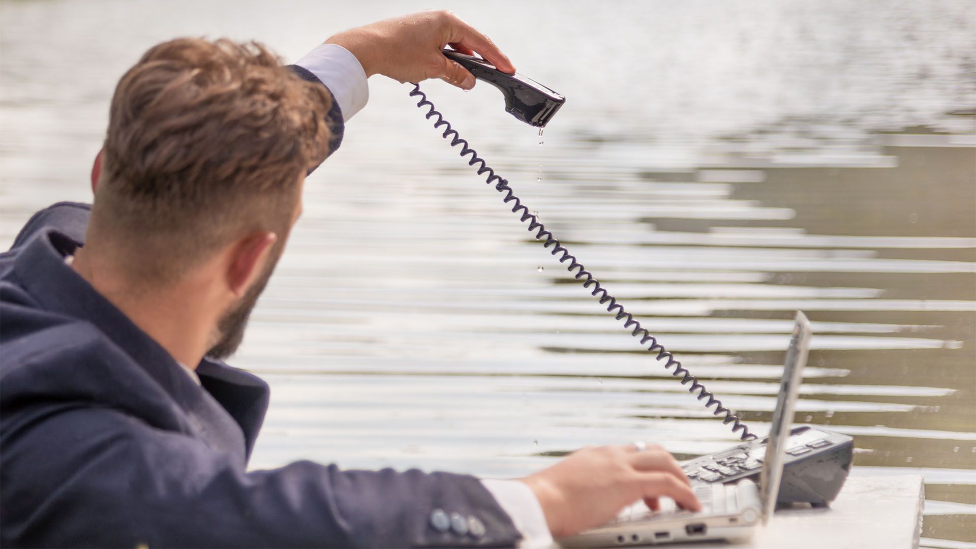 Homem estendendo a mão para a água com o fio do telefone, digitando em um laptop. Fundo desfocado com água.