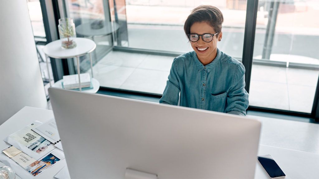 Pessoa sorrindo enquanto trabalha em um computador em uma mesa perto de uma janela.