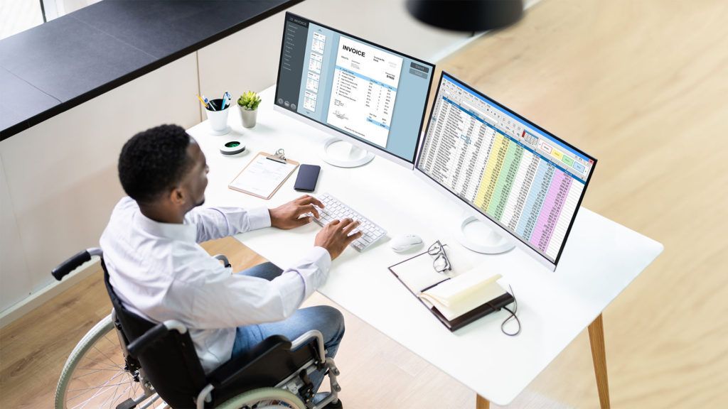 Uma pessoa em cadeira de rodas trabalha em um computador com dois monitores, analisando planilhas em uma mesa.