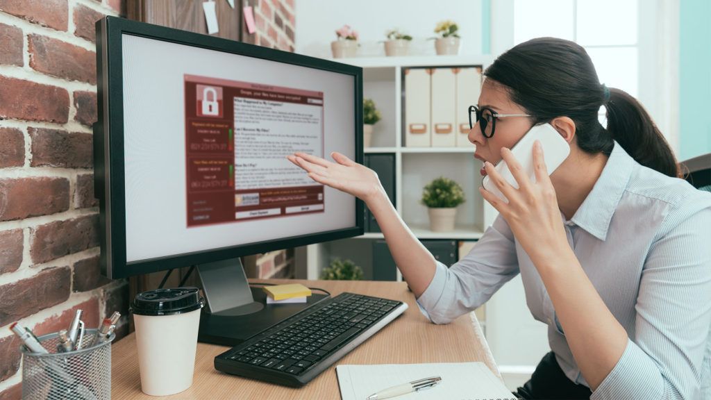 Uma mulher ao telefone gesticula em direção à tela de um computador com uma interface em tons de vermelho, em um ambiente de escritório.