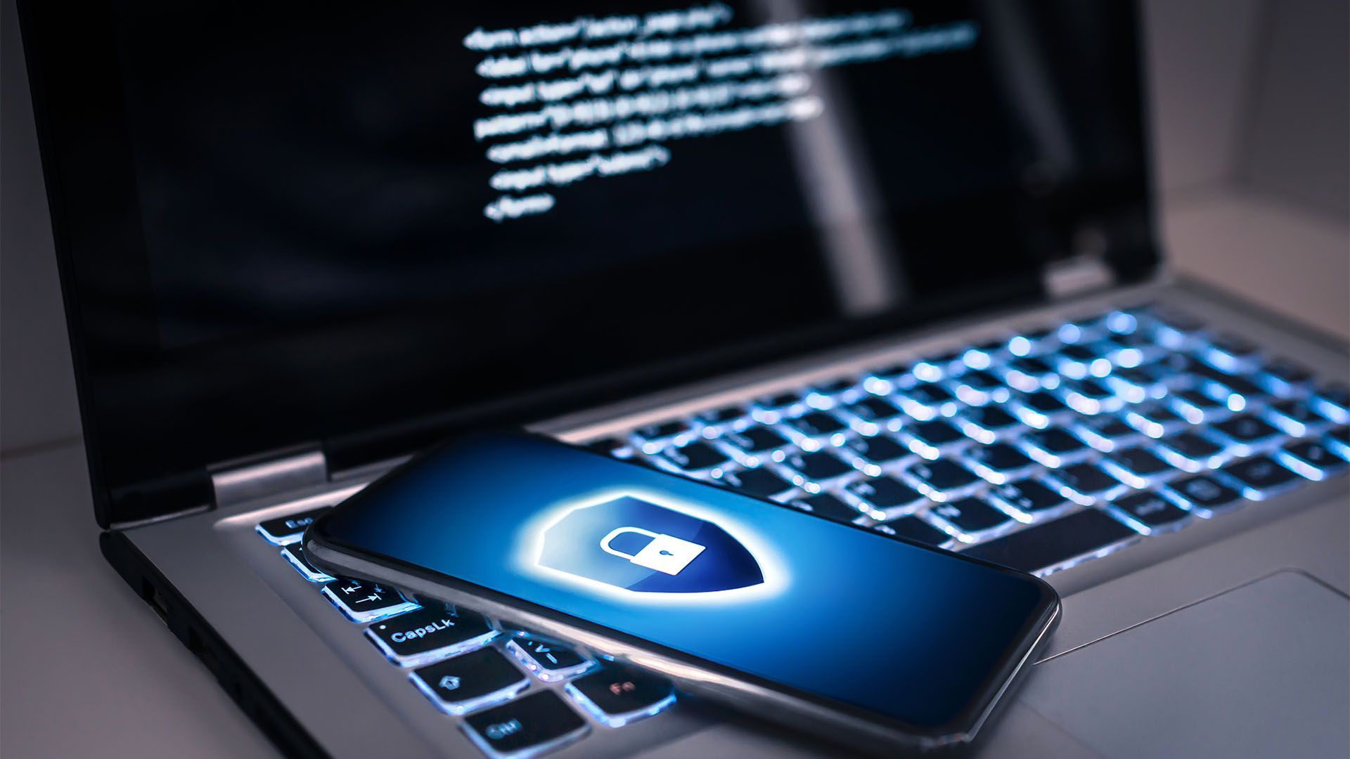 Laptop e smartphone com ícone de escudo de segurança, teclado iluminado e código na tela.