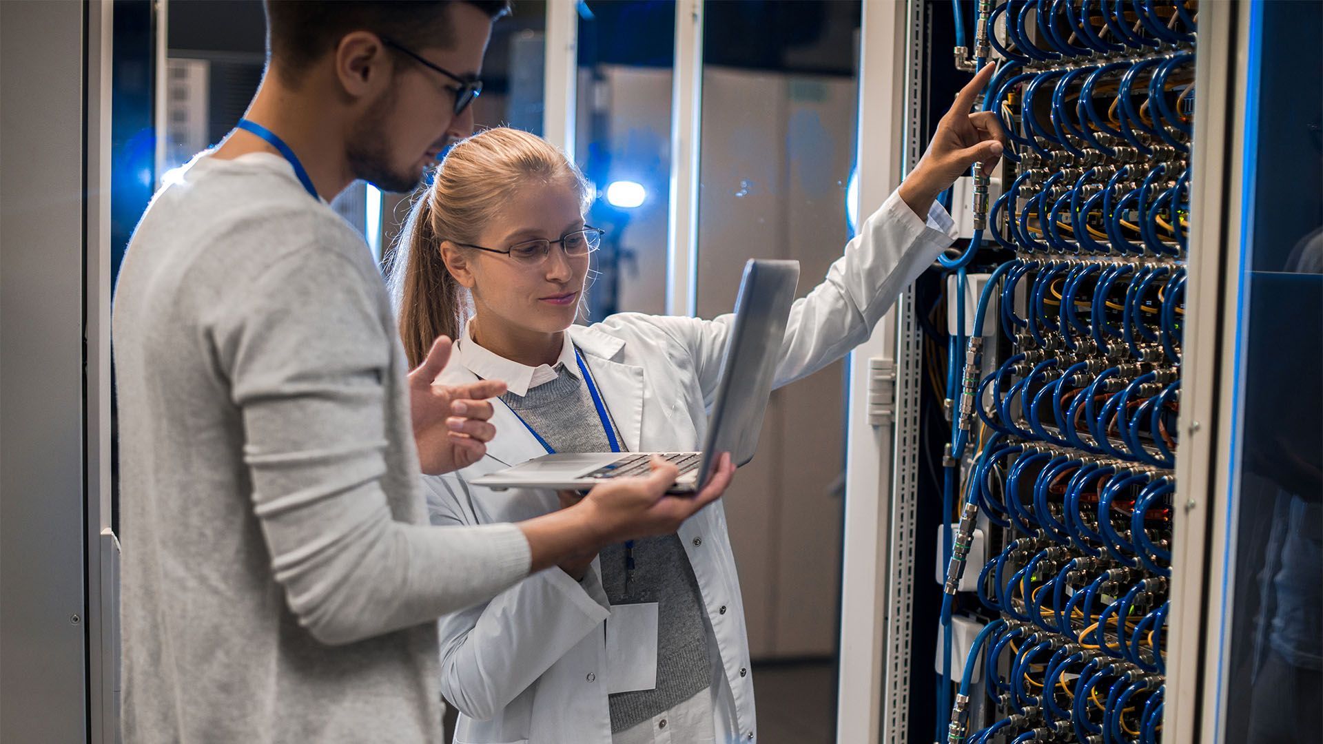 Duas pessoas examinam racks de servidores em um centro de dados; a mulher aponta e segura um laptop.