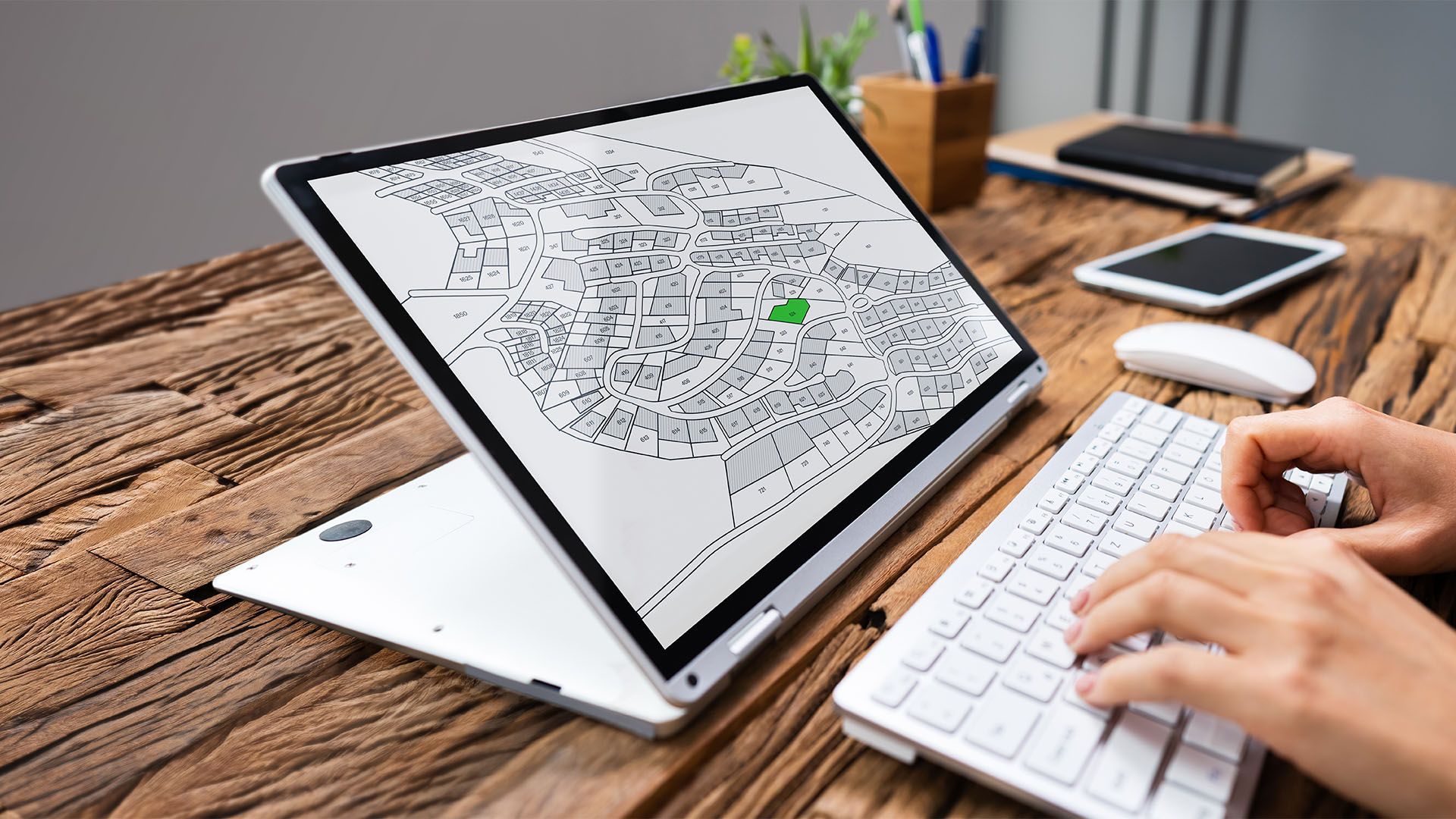 Pessoa digitando em um teclado branco, visualizando um mapa na tela de um laptop sobre uma mesa de madeira.