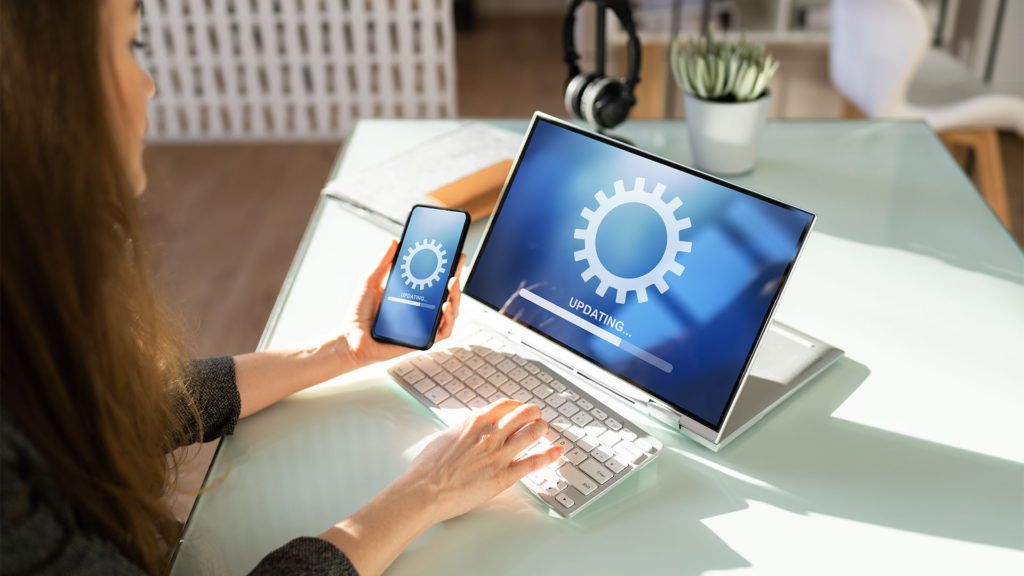 Mulher atualizando seus dispositivos: um laptop e um smartphone, com um ícone de engrenagem e uma barra de progresso em suas telas, em ambiente interno.