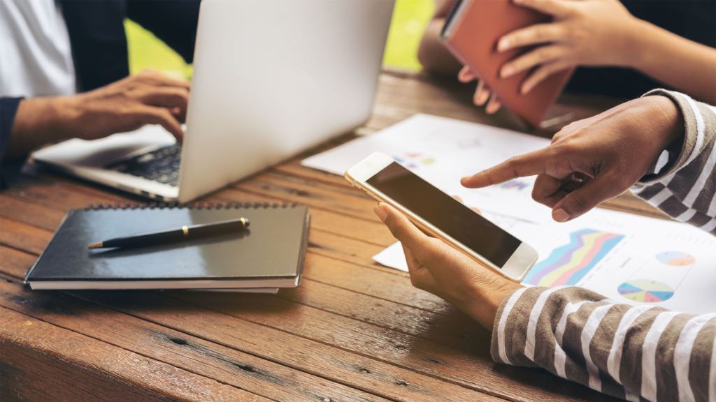 Mãos apontando para um tablet, um laptop e papéis sobre uma mesa de madeira, discutindo dados.
