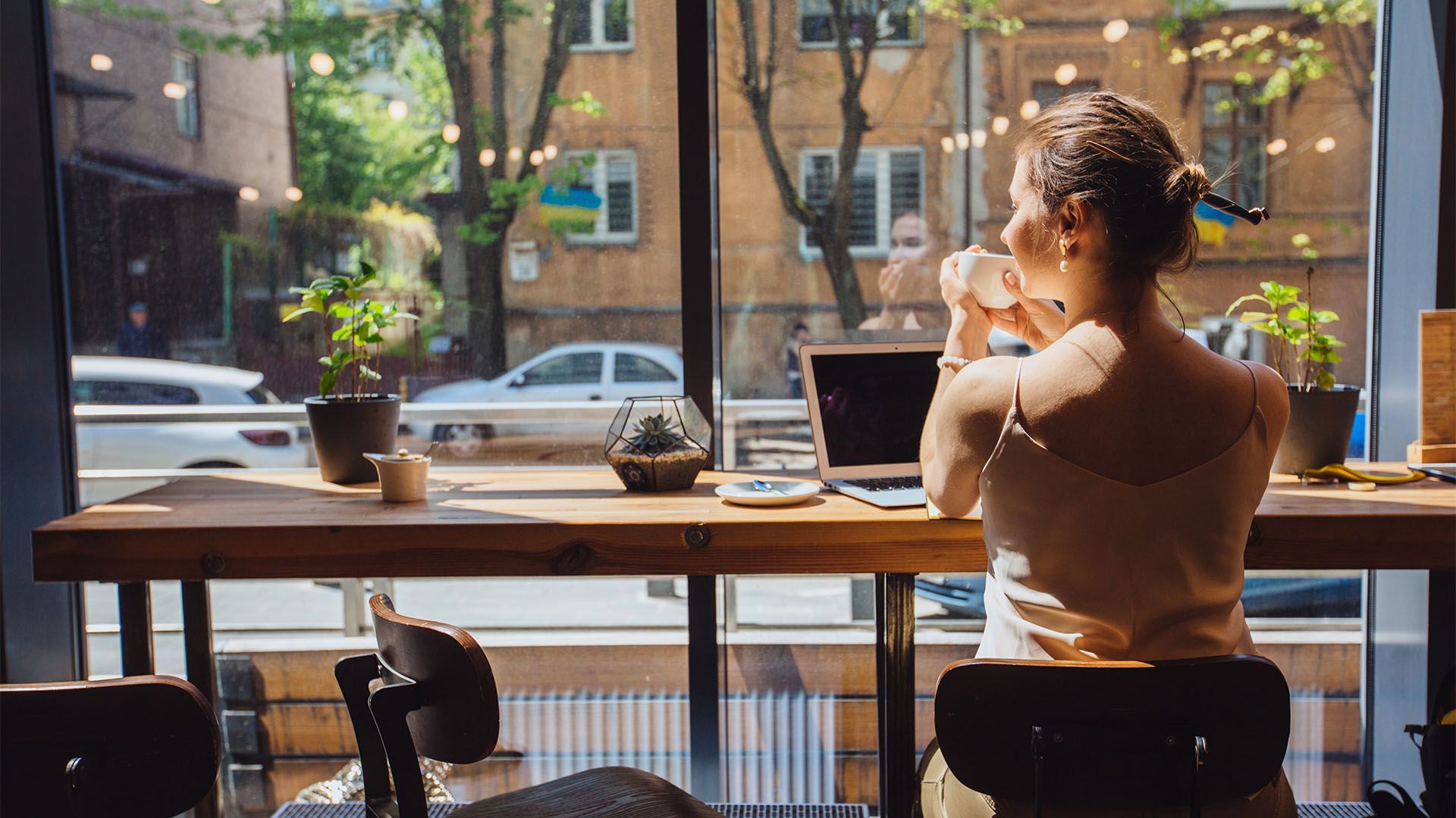 Mulher sentada à mesa perto da janela em um café, olhando para fora enquanto bebe de uma caneca com um laptop à sua frente.