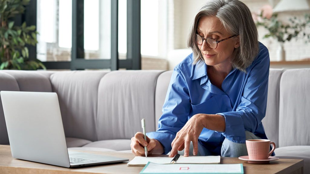 Mulher de cabelos grisalhos e óculos, escrevendo em um caderno enquanto usa um laptop sentada em um sofá, dentro de casa.