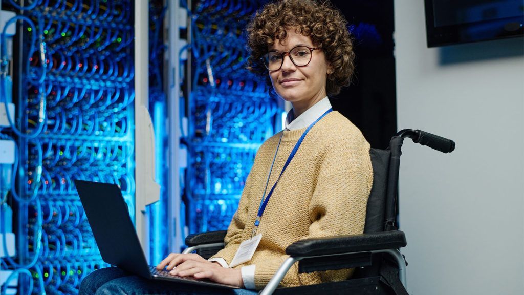 Pessoa em cadeira de rodas usando um laptop em uma sala de servidores. Ela está sorrindo.