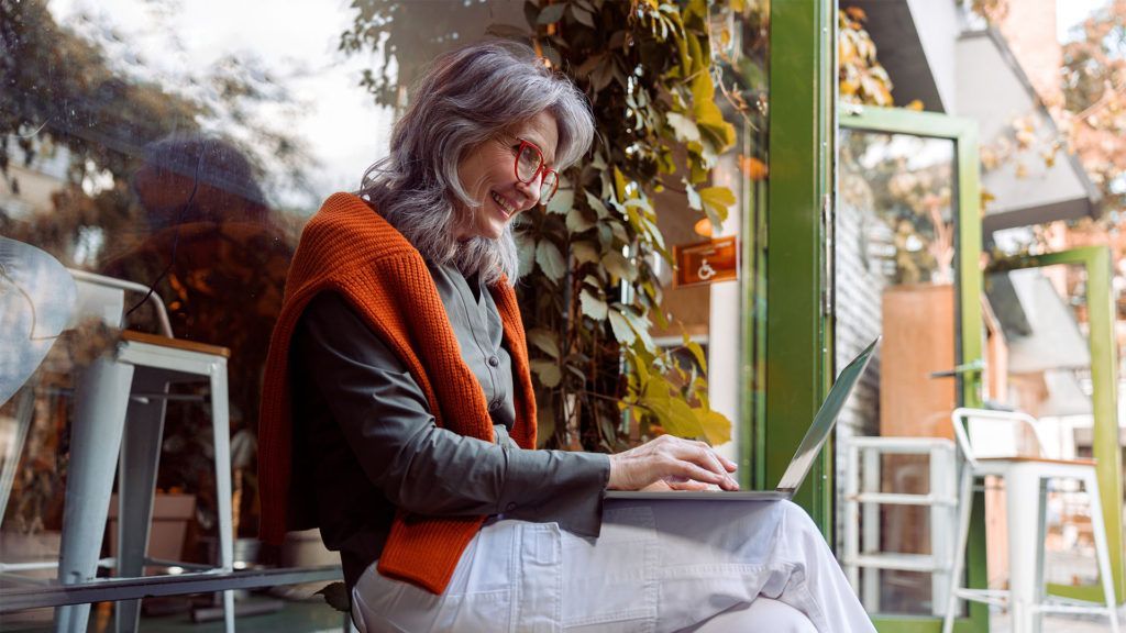 Mulher de cabelos grisalhos, vestindo um suéter laranja, trabalhando em um laptop em um café com vista para a janela.