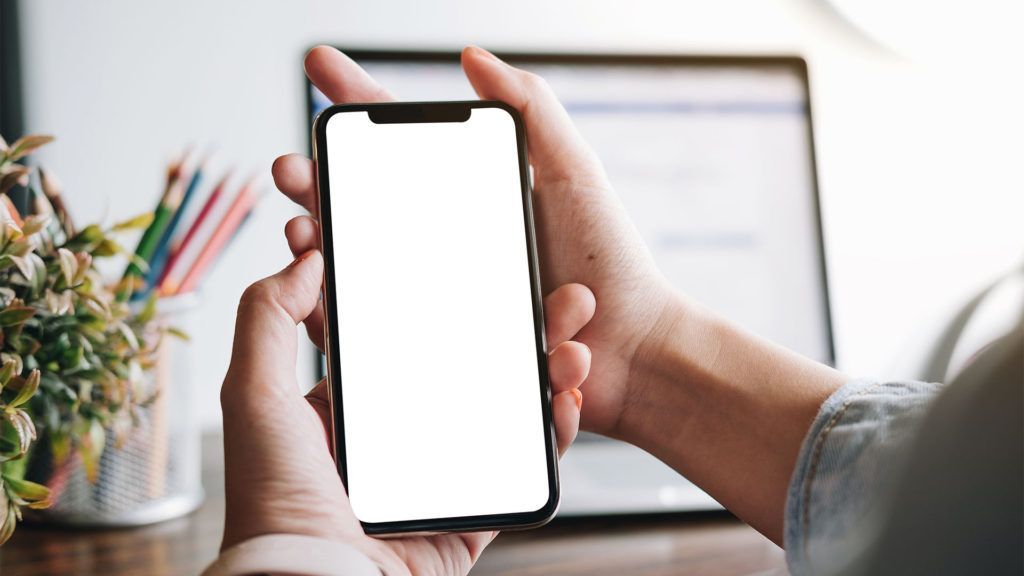 Pessoa segurando um smartphone com a tela completamente branca, tendo como fundo desfocado uma mesa e um laptop.