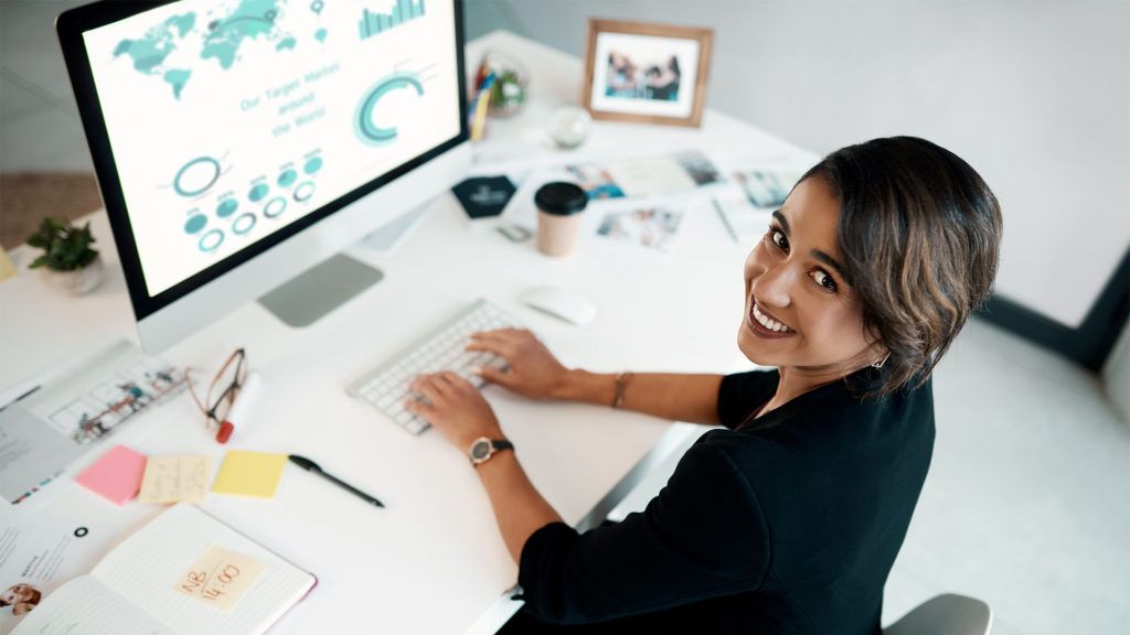 Mulher trabalhando no computador, sorrindo para a câmera em um escritório moderno.