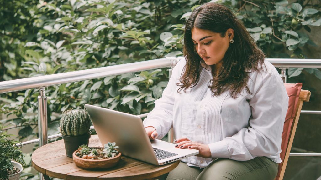 Mulher na varanda, digitando em um laptop. Mesa pequena com plantas e folhagem verde ao fundo.