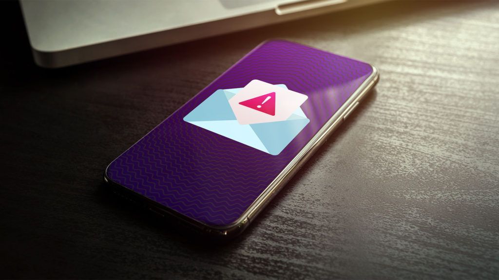 Smartphone exibindo um ícone de e-mail com um ponto de exclamação, ao lado de um laptop sobre uma superfície escura.