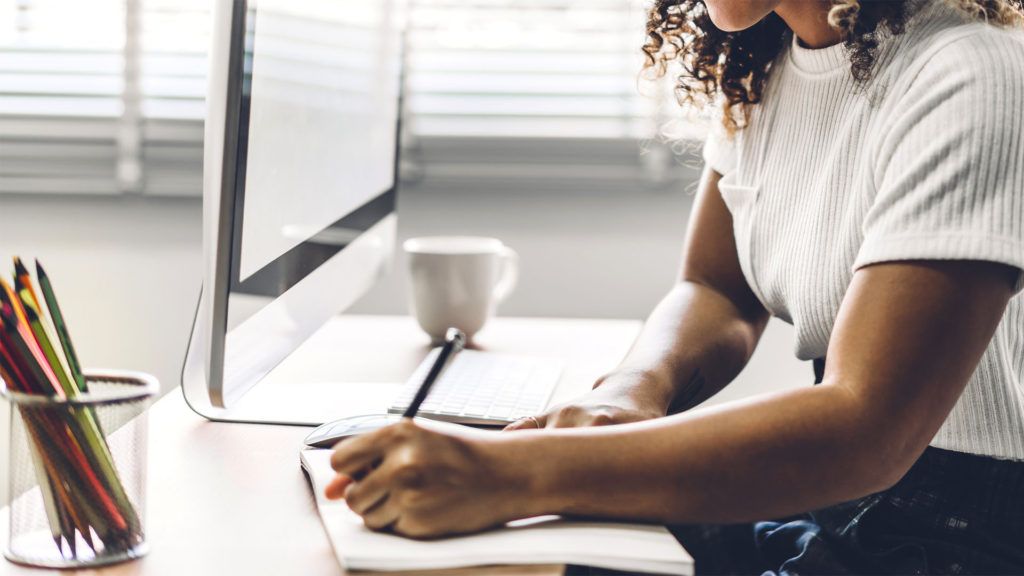 Mulher de cabelos cacheados escrevendo em um caderno em uma mesa com computador e lápis.