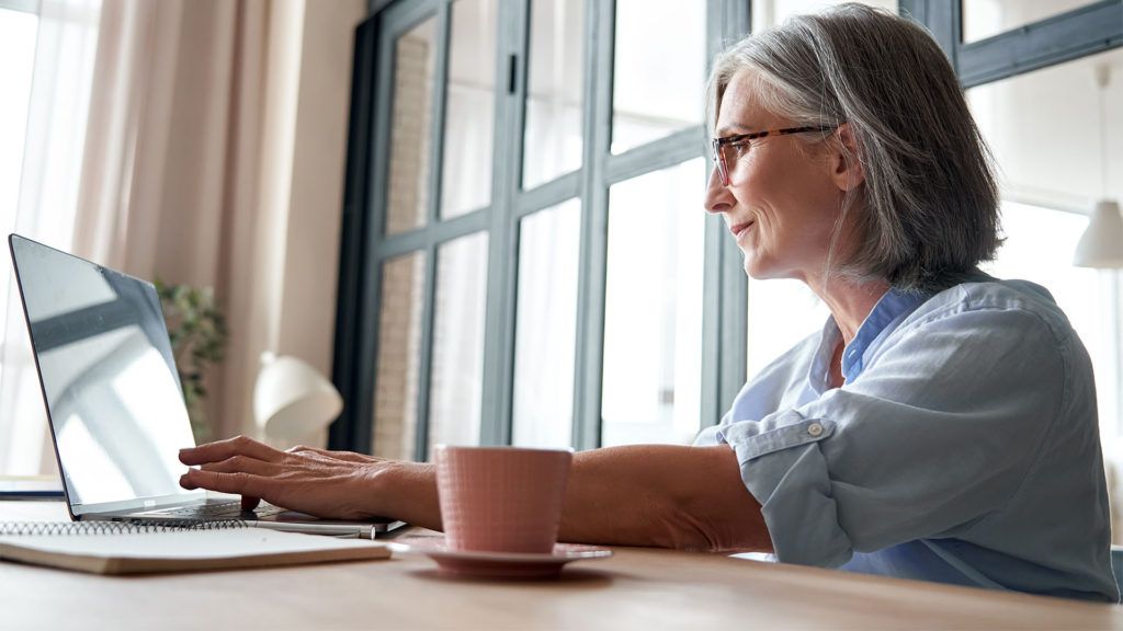 Mulher de cabelos grisalhos digitando em um laptop em uma mesa perto de uma janela, com uma xícara de café por perto.